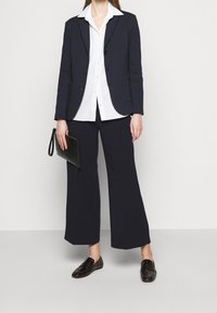 Kobieta w granatowej marynarce, białej koszuli z guzikami, szerokich granatowych spodniach, ciemnych mokasynach, trzymająca czarną kopertówkę z paskiem na nadgarstek.