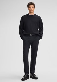 Maglione nero a coste con un collo alto, abbinato a pantaloni scuri e scarpe nere stringate, in piedi su uno sfondo grigio chiaro.