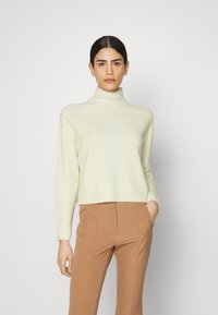 Helle cremefarbene Rollkragensweater, mit gerippten Kanten, weiche Textur, kombiniert mit hellbraunen, hoch taillierten Hosen, sitzt auf der Hüfte, enganliegende Silhouette.