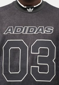 Crna rebrasta Adidas majica kratkih rukava s bijelim logotipom "ADIDAS" i grafikonom "03". Ima crni i bijeli rebrasti ovratnik. Teksturirani materijal.