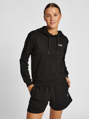 Jonge vrouw met een zwarte hoodie en bijpassende korte broek, staande met handen in de zakken tegen een effen achtergrond.