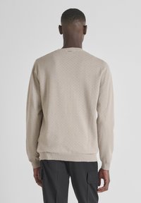Maglione beige con texture, collo a giro e polsini a costine, caratterizzato da un motivo geometrico sul retro. Abbinato a pantaloni cargo scuri.