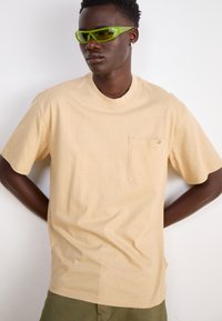 Tricou bej cu mânecă scurtă, dotat cu un buzunar și un mic logo, realizat dintr-un material moale. Asortat cu ochelari de soare verzi strălucitori.