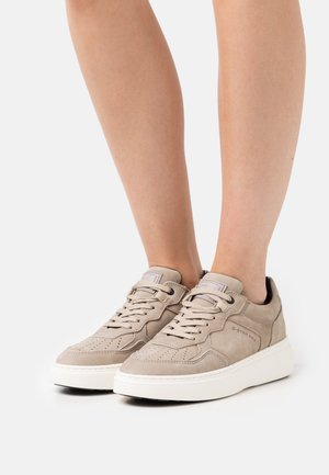 Beige suede sneakers with a smooth finish, featuring perforations, flat laces, and a white rubber sole. Brand logo visible on the side.