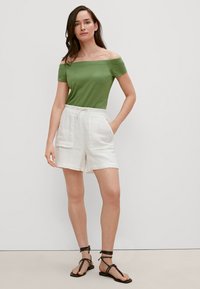 Grünes Off-Shoulder-Top, kombiniert mit weißen Leinen-Shorts mit vorderen Taschen und abgerundet mit schwarzen Riemchensandalen.