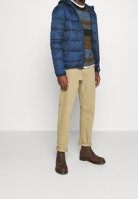 Doudoune bleue avec capuche, pull en maille à motifs verts, bruns et crème, pantalon beige retroussé et bottines marron foncé.