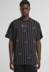 Mann trägt ein schwarzes übergroßes T-Shirt mit kurzen Ärmeln, dünnen weißen vertikalen Streifen und weißen abstrakten Linienmustern vor einem einfarbigen Hintergrund.