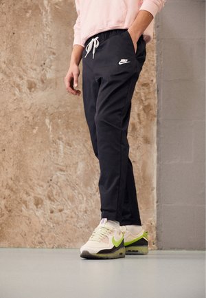 Osoba w czarnych spodniach dresowych Nike i białozielonych butach Nike, stojąca przy teksturowanej beżowo-szarej ścianie.