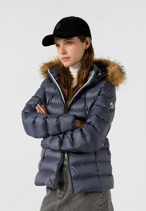 Marineblauwe pufferjack met een capuchon en faux fur rand, ritssluiting, geribbelde manchetten en een logo patch, gecombineerd met een zwarte pet.