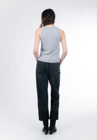 Mujer de pie con las manos en los bolsillos traseros, vistiendo una camiseta sin mangas azul claro, pantalones anchos de mezclilla oscuros con puños y botas negras de tacón.
