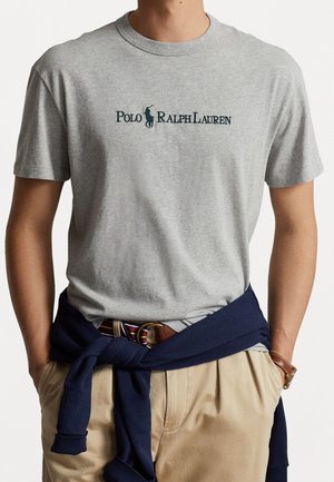 Graues Baumwoll-T-Shirt mit Rundhalsausschnitt und einem dunkelgrünen, bestickten "Polo Ralph Lauren"-Logo. Kombiniert mit beigefarbenen Hosen und einem marineblauen Pullover, der um die Taille gebunden ist.