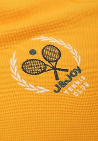 Geel getextureerde stof met een geborduurd ontwerp. Bevat twee tennisrackets en een cirkelvormig embleem, omringd door witte lauriertakken.