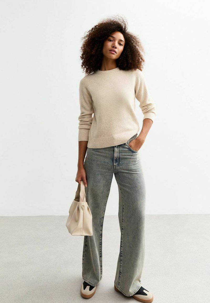 Pull beige à col rond, associé à un jean décontracté à jambes larges en denim clair. Tenant un petit sac à main structuré beige.
