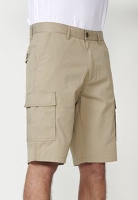Pantaloni scurți cargo bej dintr-un material ușor, cu două buzunare laterale mari și un buzunar cu fermoar pe partea dreaptă. Talie standard și bucle pentru brâu.