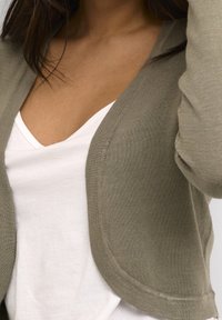 Cardigan vert olive clair en tissu tricoté doux, avec une ouverture arrondie sur le devant et une texture côtelée le long des bords. Chemise blanche à col en V en dessous.