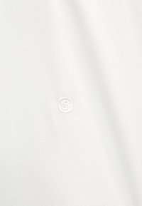 Tissu blanc avec un logo circulaire brodé subtil comportant la lettre « S » au centre.