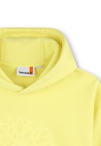 Sudadera de algodón amarilla con un bolsillo frontal, una capucha texturizada y un gran logo de copo de nieve en relieve en la parte delantera. Etiqueta de la marca visible en el interior.