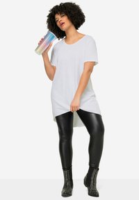 Witte, korte mouwen T-shirt, oversized pasvorm, gecombineerd met zwarte, glanzende leggings. Model houdt een meerkleurige, gestructureerde mok vast. Zwarte enkellaarzen met studs.