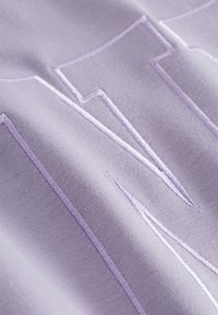 Lavendel stof met verhoogde borduursels van gestileerde letters. Gladde textuur met duidelijke stiklijnen die het ontwerp accentueren.