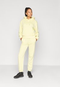Ανοιχτό κίτρινο φούτερ με κουκούλα, κορδόνια και τσέπη καγκουρό, συνδυασμένο με αντίστοιχο παντελόνι φόρμας. Το μοντέλο φοράει μαύρα αθλητικά παπούτσια. Απλός σχεδιασμός, άνετο ύφασμα.