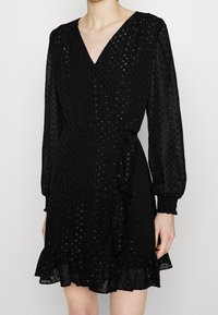 Svart omlottklänning med texturerade prickar, V-ringning, långa ärmar med smockade manschetter och volangdetalj i fållen. Tillverkad i ett lättviktsmaterial.