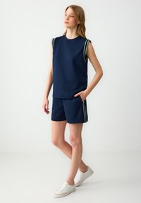 Top senza maniche blu navy con strisce sulle spalle verdi e bianche, abbinato a pantaloni corti blu navy con tasche laterali e scarpe da ginnastica bianche.