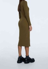 Robe midi en tricot côtelé vert olive avec des manches longues, silhouette ajustée, fente sur le côté, assortie à des bottines noires chunky et des chaussettes grises.