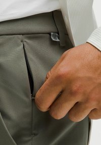 Zelene hlače od glatke tkanine, s bočnom preklopnom džepom i sivom oznakom. Ruka je djelomično vidljiva, držeći patentni zatvarač.