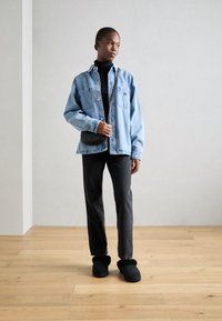 Femme portant une veste en denim bleu clair sur un col roulé noir, un pantalon noir, des chaussures noires à enfiler, et tenant un petit sac croisé noir.