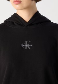 Čierna mikina s kapucňou vyrobená z mäkkej bavlnenej tkaniny, s predným logom "Calvin Klein" a iniciálami "cK" v sivej výšivke.