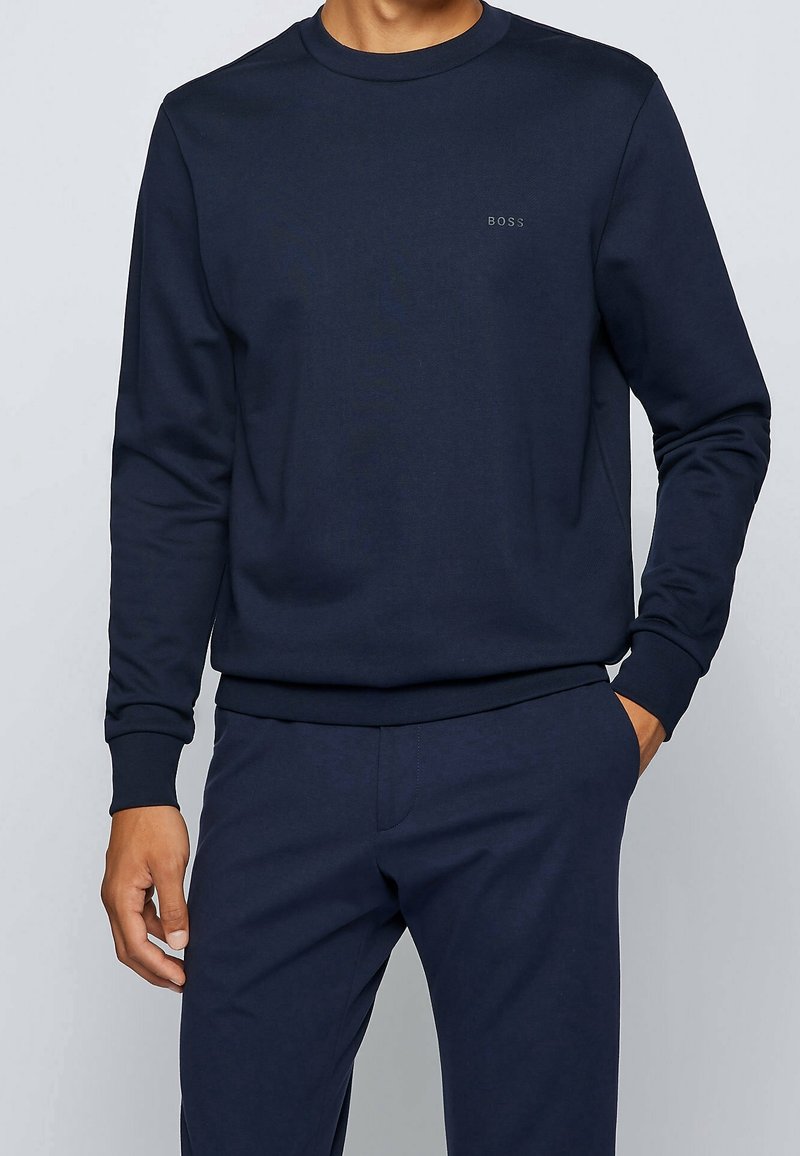 Mörkblå sweatshirt gjord av bomullsblandning, med långa ärmar, ribbade muddar och en liten logotyp på bröstet. Slät, mjuk textur.