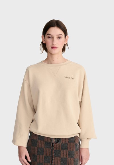 Jeune femme aux cheveux courts et foncés portant un sweat beige avec le texte "HUG ME" et une jupe à carreaux marron et noire.