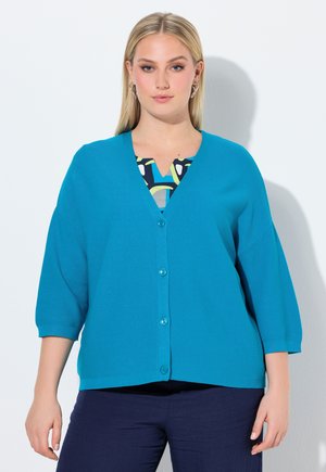 Donna con lunghi capelli biondi che indossa un cardigan verde acqua abbottonato sopra una maglia fantasia, abbinato a pantaloni blu scuro, in piedi su sfondo bianco.