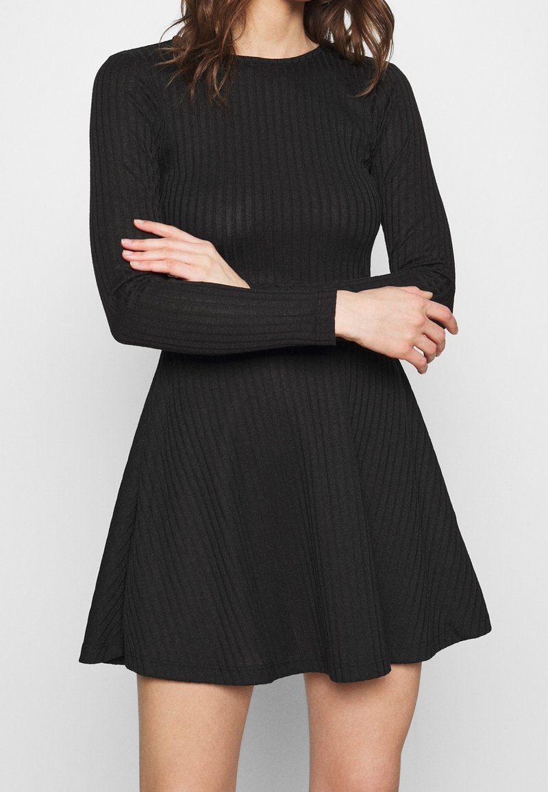 Femme portant une robe noire côtelée à manches longues, debout les bras croisés sur la poitrine devant un fond uni.