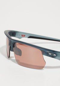 Sonnenbrille mit einem dunkelblauen Kunststoffrahmen, umschließendem Design und rosafarbenen Gläsern. Merkmale sind die große Linsenform und die seitlichen Blenden.