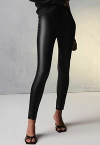Leggings in finta pelle nera con vita alta, texture liscia e cuciture a contrasto. Abbinati a sandali neri con tacco e cinturini.