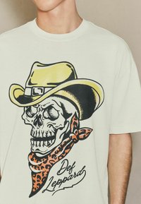 Kreemivärvi T-särk, millel on graafika koljust, kes kannab kollast cowboy-mütsi ja oranži leopardimustriga sall, koos tekstiga "Def Leppard".