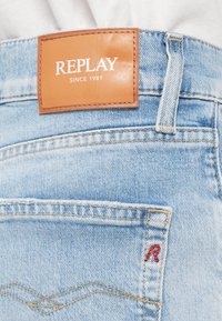 Ανοιχτό μπλε τζιν παντελόνι με καφέ δερμάτινη ετικέτα που γράφει "REPLAY SINCE 1981" και κόκκινο κεντημένο "R" στην πίσω τσέπη.