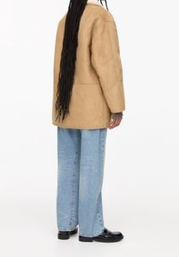 Tanfarvet ruskindjakke med afslappet pasform, sænkede skuldre og syningsdetaljer, kombineret med lyseblå vidde-bukser og sorte sko.