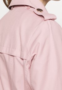 Gros plan d'une personne portant un trench-coat rose clair montrant la partie arrière de l'épaule avec un détail d'épaulette boutonnée.