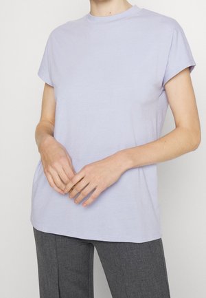 Person trägt ein hellviolettes T-Shirt mit kurzen Ärmeln und dunkelgraue Hose, Hände entspannt und überlappend auf Taillenhöhe.