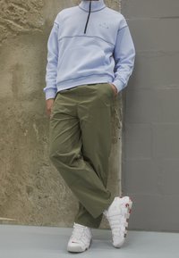 Svetle modrá mikina na zip s přední kapsou, v kombinaci s olivově zelenými kalhotami a bílými teniskami s červenými akcenty, na pozadí texturované zdi.