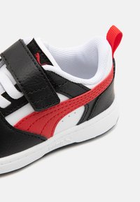 Les baskets noir et rouge présentent un supérieur en cuir lisse, une doublure en mesh blanche, une sangle en Velcro et une semelle en caoutchouc texturée.