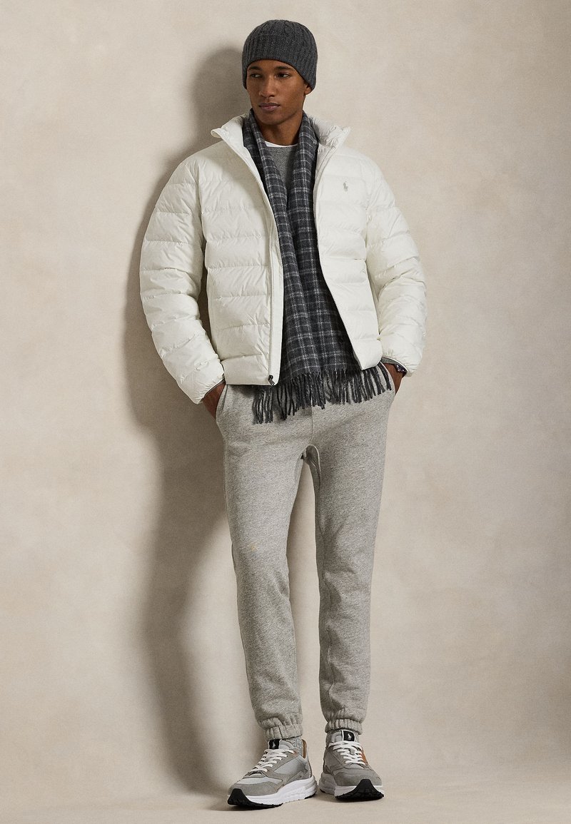 Veste matelassée blanche, écharpe grise à motif à carreaux, pantalon de jogging gris, et baskets gris/blanc avec accents orange. Un bonnet tricoté complète la tenue.