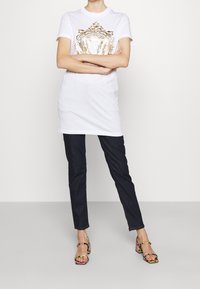 Biała koszulka z krótkim rękawem z złotym nadrukiem na środku, w połączeniu z ciemnoniebieskimi spodniami w slim fit oraz kolorowymi sandałami na paski.