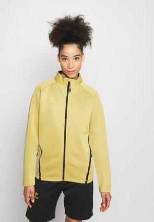 Geel fleecejack met een hoge kraag, zwarte rits, twee met rits afgesloten zijzakken en contrasterende stiksels. Gekozen in combinatie met zwarte shorts.
