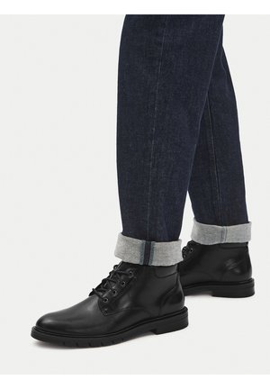 Schwarze Lederstiefel mit glattem Finish, Schnürdesign und Gummisohle, kombiniert mit dunkelblauen Jeans, die einen umgeschlagenen Saum haben.