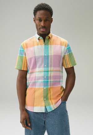 Mand med kort krøllet hår iført flerfarvet pastelternet kortærmet skjorte med knapper og blå jeans, stående med den ene hånd i lommen.