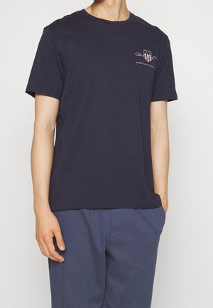 Mann trägt ein marineblaues Kurzarm-T-Shirt mit kleinem weißem und rotem Grafiklogo auf der Brust, kombiniert mit blauen Hosen, Hände entspannt.