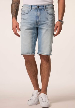 Hellblaue Jeansshorts, getragen von einem Mann mit tätowiertem linken Arm, weißen Turnschuhen und einer Armbanduhr, stehend vor einem schlichten Hintergrund.